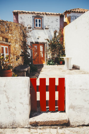 Close-up of a vivid red wooden gate set in a white stucco wall, leading into a charming Mediterranean courtyard with stone walls, potted plants, and a rustic double door in soft sunlightの写真素材