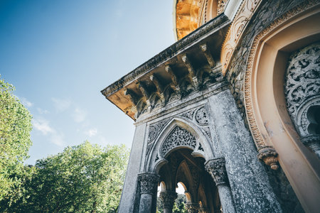 Low-angle architectural detail of a grand historical structure with ornate stone arches, intricate carvings, and Moorish influences, set against a bright blue sky and lush green treesの写真素材