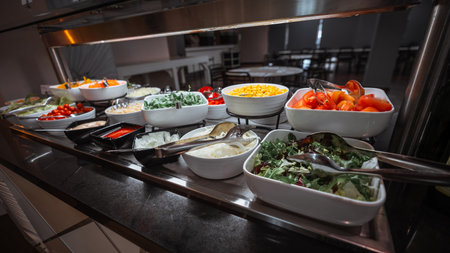 Colorful self-service salad buffet with a variety of fresh vegetables including tomatoes, corn, onions, lettuce, arugula, cherry tomatoes, and sauces, displayed in white bowls under warm lightingの写真素材
