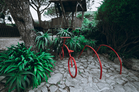 Artistic red bicycle-shaped metal bike rack surrounded by lush green tropical plants and trees, placed on a cobblestone path near a rustic wooden treehouse in a shaded forest setting; wide-angle shotの写真素材