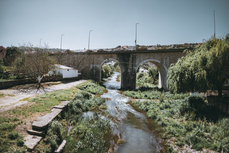 Scenic view of a historic stone bridge with multiple arches spanning a tranquil river, surrounded by lush greenery and a rustic white house under clear blue skies in a peaceful couの写真素材