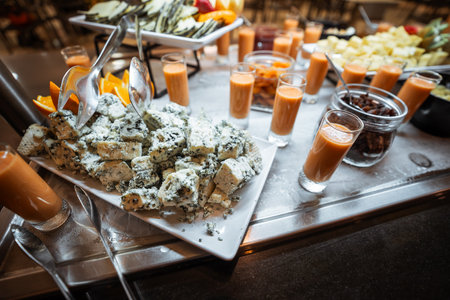 Gourmet buffet table with a close-up of creamy blue cheese cubes garnished with orange slices, surrounded by small glasses of fresh smoothie, dried fruit, and assorted cheese selectionsの写真素材