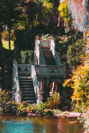 Elegant stone staircase with gothic-style balustrades rises symmetrically from a garden pond,...