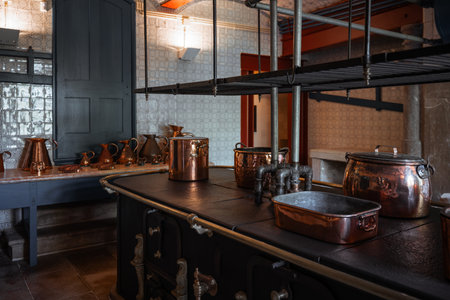 Low-key interior of a historic kitchen featuring a vintage cast iron stove with aged copper pots and pans, surrounded by tiled walls and traditional dark cabinetry under moody lightingの写真素材