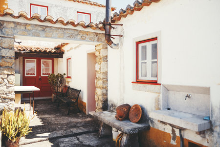 Traditional rustic courtyard in Aldeia da Mata Pequena, Portugal, featuring whitewashed walls, red window frames, stone details, ceramic pots, and an old outdoor sink bathed in sunlightの写真素材