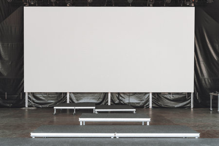 Large blank LED screen or digital billboard mockup set up on an indoor stage with empty risers and black curtains, perfect for inserting custom visuals, event branding, or advertising contentの写真素材