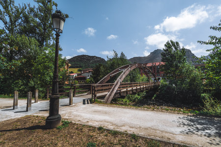 Wooden arch pedestrian bridge in Las Arenas, Cabrales, surrounded by lush greenery, traditional Spanish buildings, and mountainous landscape under a clear blue summer sky, peaceful village vibeの写真素材