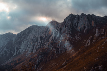 Dramatic rocky slopes of Picos de Europa under moody evening clouds, with a single ethereal sunbeam breaking through the sky and illuminating a cliff face, creating a mystical, cinematic atmosphereの写真素材