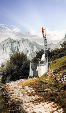 Remote telecommunications tower with orange antenna and satellite dish, enclosed by a metal fence and set on a mountain slope with rugged cliffs and dramatic sky in the backgroundの写真素材