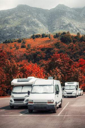 Several camper vans parked in an autumn forest lot at the foot of dramatic mountains, surrounded by vibrant red foliage and misty peaks, capturing the spirit of outdoor travel and freedomの写真素材