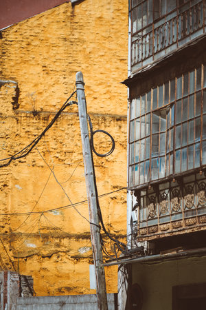 Rustic urban composition in Tineo, featuring a weathered wooden utility pole with tangled wires set against a vibrant textured yellow wall and ornate vintage ironwork on an old glass facadeの写真素材