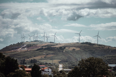 Panoramic view of a wind farm with multiple wind turbines spread across a rolling green hill under a dramatic cloudy sky, with scattered rural houses and trees in the foregroundの写真素材
