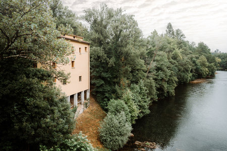 Secluded building with warm yellow facade nestled in dense green forest on the riverbank in Asturias, Spain, surrounded by lush trees and reflecting water under a soft overcast skyの写真素材