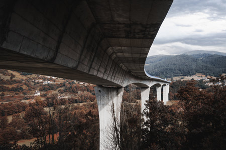 Dramatic low-angle view of a sweeping modern concrete bridge curving across an autumnal valley with forested hills and scattered rural houses under a moody overcast skyの写真素材