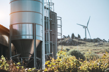 Shiny metal grain silos with vertical ladders on a rural farm, set against a grassy hillside and a wind turbine in the distance under clear blue sky with soft sunlight flare; Asturias, Spainの写真素材