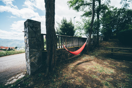Bright red hammock hanging between trees in a shaded corner of a small park, near a stone fence with mountain views and village rooftops visible in the background under partly cloudy sky; wide-angleの写真素材