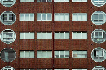 Symmetrical facade of a red-tiled apartment building with a grid of rectangular windows and unique round balcony enclosures on both sides, showcasing modernist residential architecture, urban patternの写真素材