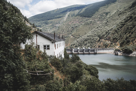 Old white building nestled in lush greenery on a hillside overlooking a reservoir and large concrete dam, surrounded by forested mountains and power lines under a partly cloudy summer skyの写真素材