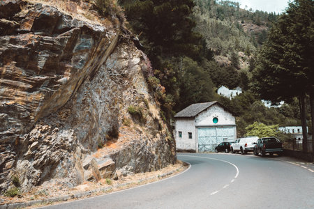 Winding mountain road curves past a rugged rock cliff and leads to a small white industrial building with a green circular window, surrounded by parked cars and dense pine forest on the hillsの写真素材