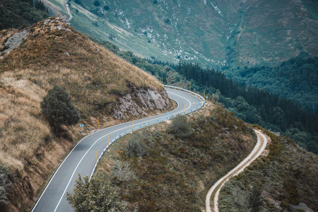 Telephoto view of a winding asphalt mountain road with safety railings, flanked by a rugged hiking trail, cutting through scenic green hills and forested slopes in a peaceful natural landscapeの写真素材