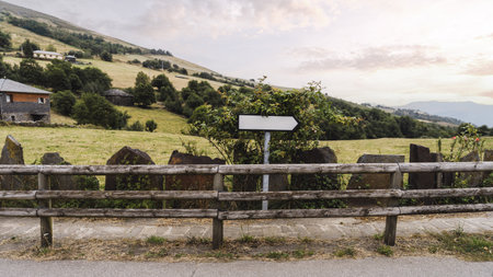 Blank arrow sign mockup in a rustic countryside setting, positioned behind a wooden fence and lush greenery, with hills, stone houses, and soft sunset light creating a warm and natural template backdropの写真素材