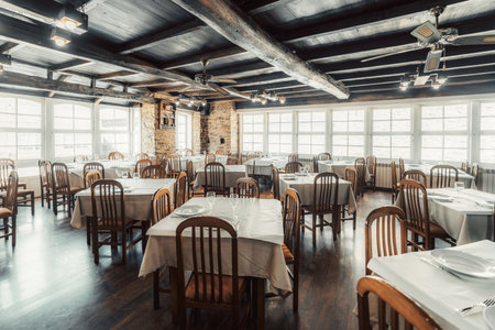 Spacious rustic-style restaurant interior with wooden beams, stone walls, and neatly arranged tables covered in white cloth, set for guests with glassware and plates, bathed in natural lightの写真素材