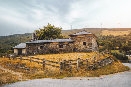 Traditional Asturian stone church with slate roof and rustic bell gable, surrounded by wooden fence on hillside meadow, with wind turbines visible on the distant ridgeの写真素材