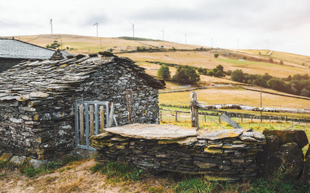 Traditional Asturian rural architecture with rustic stone house built from slate and local rock masonry, wooden gate and fence, set in countryside hills with wind turbines on horizonの写真素材