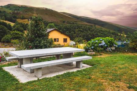 Scenic countryside garden with stone picnic table and benches, colorful flowers, and rustic yellow house surrounded by lush green hills under dramatic sunset skyの写真素材