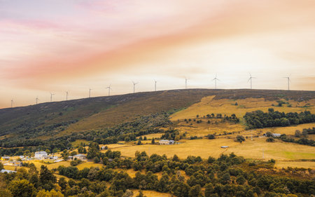 Scenic autumn countryside landscape with rolling hills, scattered houses, golden fields, and a row of modern wind turbines along the ridge under vibrant sunset skyの写真素材