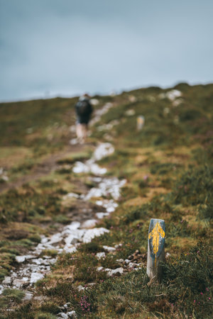 Yellow arrow trail marker on wooden post along rocky mountain path of Camino Primitivo, with blurred pilgrim hiker walking uphill on Camino de Santiago route; telephoto shotの写真素材