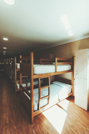 Spacious hostel dormitory interior with multiple wooden bunk beds, clean white bedding, lockers and natural sunlight creating a warm welcoming atmosphere for travellers; wide-angle vertical shotの写真素材
