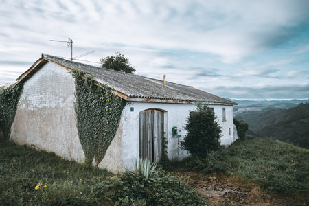 Old rural farmhouse with weathered white walls, ivy climbing facade, wooden doors, overgrown plants and scenic mountain valley view under cloudy blue sky in Asturias countrysideの写真素材