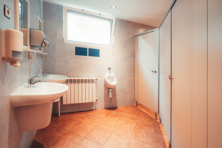 Modern public restroom with clean sinks, mirrors, soap dispensers, urinal, tiled floor, bright lighting and toilet stalls, designed for hygiene and functionality in shared facilitiesの写真素材