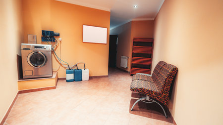Modern laundry room with industrial washing machine, colorful patterned chair, orange walls, tiled floor, plastic detergent canisters and shelf stacked with clean folded towelsの写真素材