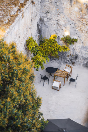Cozy outdoor patio with a rustic stone wall, wooden and metal chairs around small tables, surrounded by green foliage creating a charming Mediterranean atmosphereの写真素材