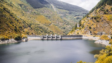 Large concrete dam across a calm mountain reservoir, surrounded by green and yellow hillsides with scattered trees and abandoned stone structures on rocky slopesの写真素材