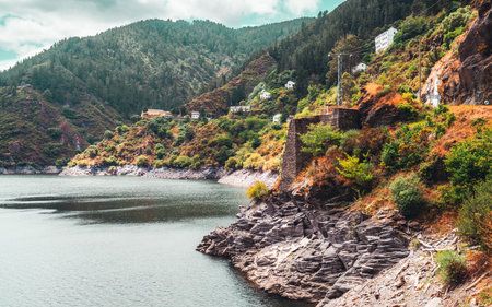 Scenic mountain reservoir with rocky shoreline, green pine-covered hills, and scattered white houses perched on slopes, creating a picturesque lakeside village viewの写真素材