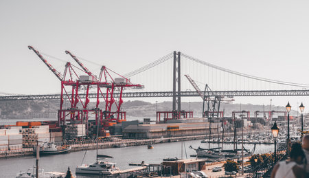 Industrial port with towering red cranes loading colorful shipping containers under a large suspension bridge, with boats and yachts docked in the foreground at the marinaの写真素材