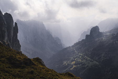 Dramatic view of Picos de Europa mountains with jagged cliffs, deep valleys, and misty atmosphere as sunlight breaks through thick clouds over the rugged autumn landscapeの写真素材
