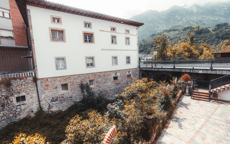 Traditional stone and stucco buildings in Cabrales, Asturias, with a scenic stone-paved courtyard, lush autumn foliage, and a bridge overlooking misty mountain peaks in the distance; wide-angle shotの写真素材