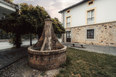 Historic stone fountain in a quiet square in Cabrales, Asturias, Spain, surrounded by cobblestone pavement, a vine-covered pergola, and traditional stone architecture at duskの写真素材