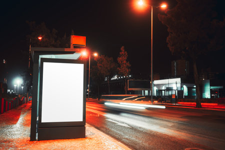 Blank illuminated billboard mockup at a bus stop on a city street at night, with long exposure light trails from passing cars and glowing street lamps creating a modern urban sceneの写真素材