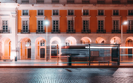Long exposure night shot of a bus stop in Lisbon with motion blur of passing vehicles, illuminated street lamps, and historic architecture with bright yellow facade in the backgroundの写真素材