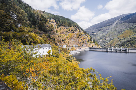Scenic autumn mountain reservoir with a dam, surrounded by lush green and yellow foliage, rocky slopes, and a white house nestled among the trees under a bright cloudy skyの写真素材