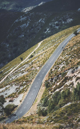 Steep winding mountain road cutting through rocky slopes and green vegetation, with protective guardrails and scenic views of rugged hillsides under natural sunlightの写真素材
