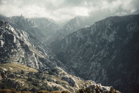 Dramatic landscape of Picos de Europa mountains with steep rocky cliffs, deep valleys, and misty clouds covering the rugged peaks, creating a breathtaking natural sceneryの写真素材