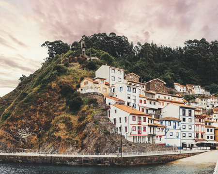 Picturesque view of colorful hillside houses in Cudillero, Asturias, Spain, perched dramatically above the harbor with lush greenery and a vibrant sunset sky in the backgroundの写真素材