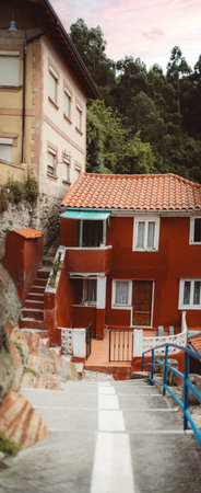 Vertical panorama of a rustic red house with tiled roof and wooden door in Cudillero, Asturias, Spain, captured from a narrow stairway with blue railings and lush greenery; shallow depth of fieldの写真素材