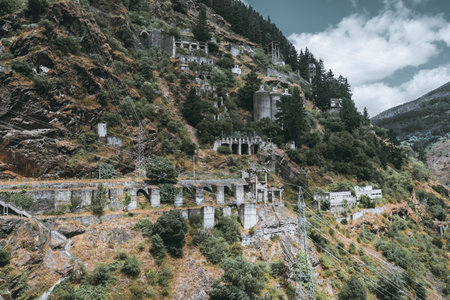 Abandoned hydroelectric complex clinging to a steep mountain, with crumbling concrete arches, silos and pylons threaded by power lines, overgrown by trees and shrubs under dramatic cloudsの写真素材